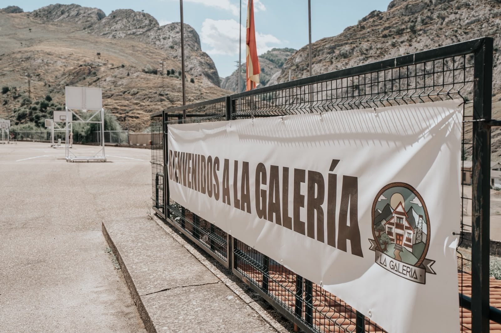 Pancarta de bienvenida a La Galería junto a las pistas de baloncesto, con las montañas y la bandera de León